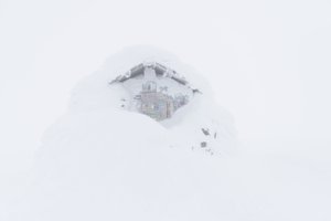 The emergency hut at the top of Ben Nevis. It’s covered in lots of snow, so only a patch of the top of the doorway is visible. There’s low visibility so it looks somewhat ghostly.