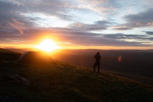 Sunrise over Whernside. Chris stands in the foreground in silhouette, taking a photo of the sun.