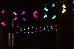 Pairs of brightly coloured lights in the shape of shoes dangle from a line suspended between two rows of houses.
