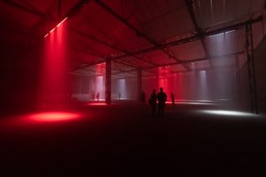 A cavernous dark warehouse. Several lights can be seen pointed at the floor like spotlights - some red and some white. Several figures can be seen silhouetted in the dark.