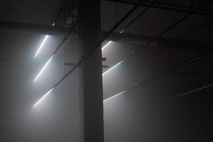 A dark scene with a single concrete pillar in the middle left. Six lines of light are evenly spaced vertically either side of the pillar. There’s fog in the room. Cables extend to and from the lines of light - reminiscent of telegraph poles.