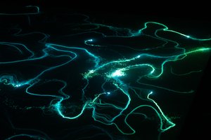 A photo from a moving light installation. A bright point light is projected on the floor, and wanders randomly around. The floor is covered in a material that glows for a period when exposed to light. The end effect is of random lines of light green and blue drawn all over the ground.