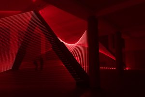 Red lasers in a dark room form lines that sweep up to a column then down again like threads.