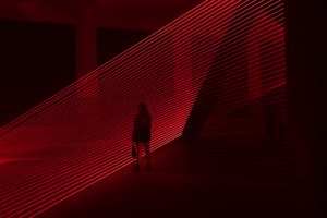 Red lasers in a dark room form even horizontal lines going from bottom left to upper right. A figure is silhouetted in front of them.
