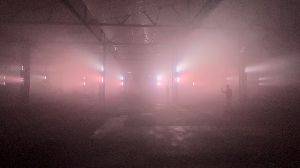 A loud audio visual video. The video is shot inside a large warehouse with bright red lights, facing the side of the warehouse strait on. As the video plays, loud rhythmic sounds can be heard. The lights pulse up the columns of the warehouse in red and white in time to the sounds.