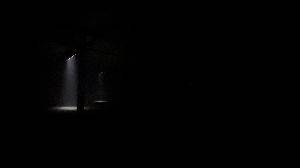 A video in a dark large warehouse. Small sections are light from above and ominous loud tones are playing. As the video progresses, lighting moves in a straight line somewhat towards the camera. At a certain point it resets back to the far room and then advances quickly towards the camera again. The lighting is timed to the music.