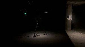 In a dark room a tripod is in the centre. On top of the tripod is a lamp and speaker connected at a pivot point. The pivot is spinning horizontally. There’s a loud clicking noise that is synchronised to the speed of the spinning.