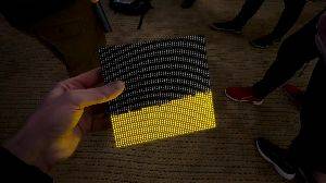 A flat square object is held in a hand. The front is completely filled with small LEDs. The lower half are lit up yellow. As the object is tilted to the side, the lit LEDs flow that way. As it is shaken the LEDs respond as if they are fluid moving about on the screen.