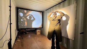 A woman in a white shipping container slowly moves her arms. In front of her a video projection slowly updates with AI images responding to her motions.