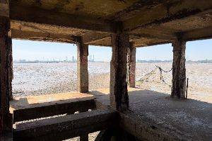 Interior photo looking outwards with exposed concrete pillars. In the distance are muddy grounds and industry.