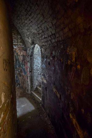 Dark photo taken in a curving corridor. The only light comes from an exit going to the right and up some stairs. Graffiti can just be made out on the walls.