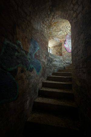 Dark photo looking up a curving stairway corridor with light at the end. There is graffiti all acros the walls.