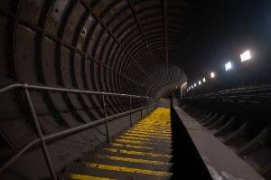 A staircase descends a dimly lit tunnel. There are regular dim lights on one side and the stair treads are painted yellow.