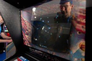 Close up photo of the screen of a vintage Asteroids arcade game  with dark black screen and white ship and asteroids. There’s strong reflections on the cover glass and the player’s face is reflected twice.
