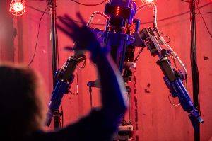 A robot with arms in the background and a human arm in the foreground. The robot is in focus.