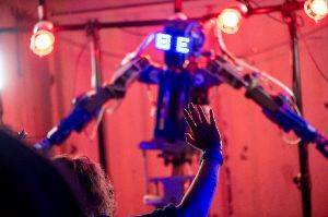 A robot with arms in the background and a human arm in the foreground. The human is in focus.