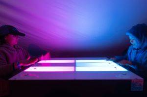 Looking side on to a flat game table that is lit up. A player is on each side.