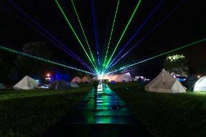 Looking straight down a path at night with white tents on either side. In the distance are lasers starting at the end of the path and going out in all directions in the sky.