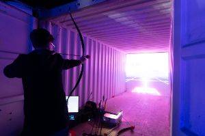Person aiming a bow and arrow in to a shipping container with large screen at the far end.