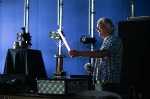 Man in a dark room holding a 2′ fluorescent tube in front of a small tesla coil. The tube is lit up and there are purple sparks from the coil to the tube.