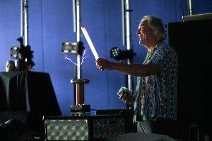 Man in a dark room holding a 2′ fluorescent tube in front of a small tesla coil. The tube is lit up and there are purple sparks from the coil to the tube.