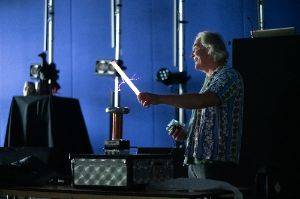 Man in a dark room holding a 2′ fluorescent tube in front of a small tesla coil. The tube is lit up and there are purple sparks from the coil to the tube.