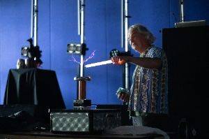 Man in a dark room holding a 2′ fluorescent tube in front of a small tesla coil. The tube is lit up and there are purple sparks from the coil to the tube.