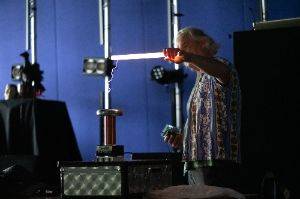 Man in a dark room holding a 2′ fluorescent tube in front of a small tesla coil. The tube is lit up and there are purple sparks from the coil to the tube.