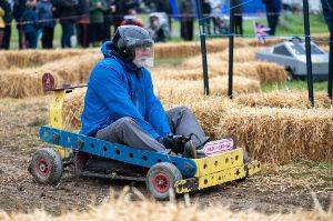 Person driving a go-kart in the shape of a Meccano car with the name ‘MEC-CAR-NO’ among hay bales as part of Wacky racers.