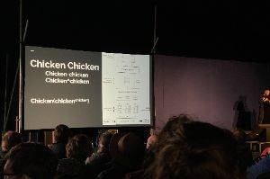 Photo of a projection screen in front of an audience. The slide has the words “Chicken Chicken, Chicken-chicken, Chicken^chicken, Chicken(chickenchicken)” and then a data flow diagram about chicken.